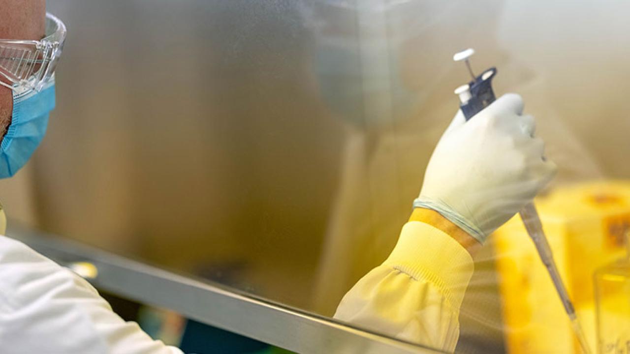 a scientist in protective gear, working behind a glass divider, using a pipette
