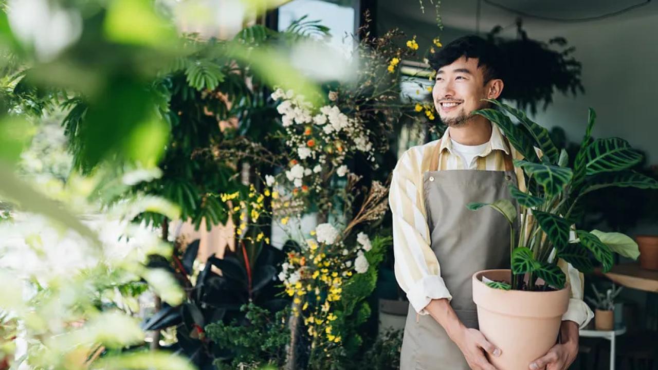 A smiling person holding a potted plant. Other plants hung around them.
