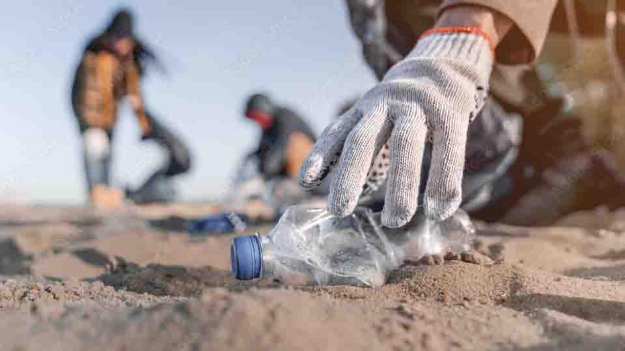 A gloved hand reaching down to pick up an empty plastic bottle on a beach. Others blurred in the background.