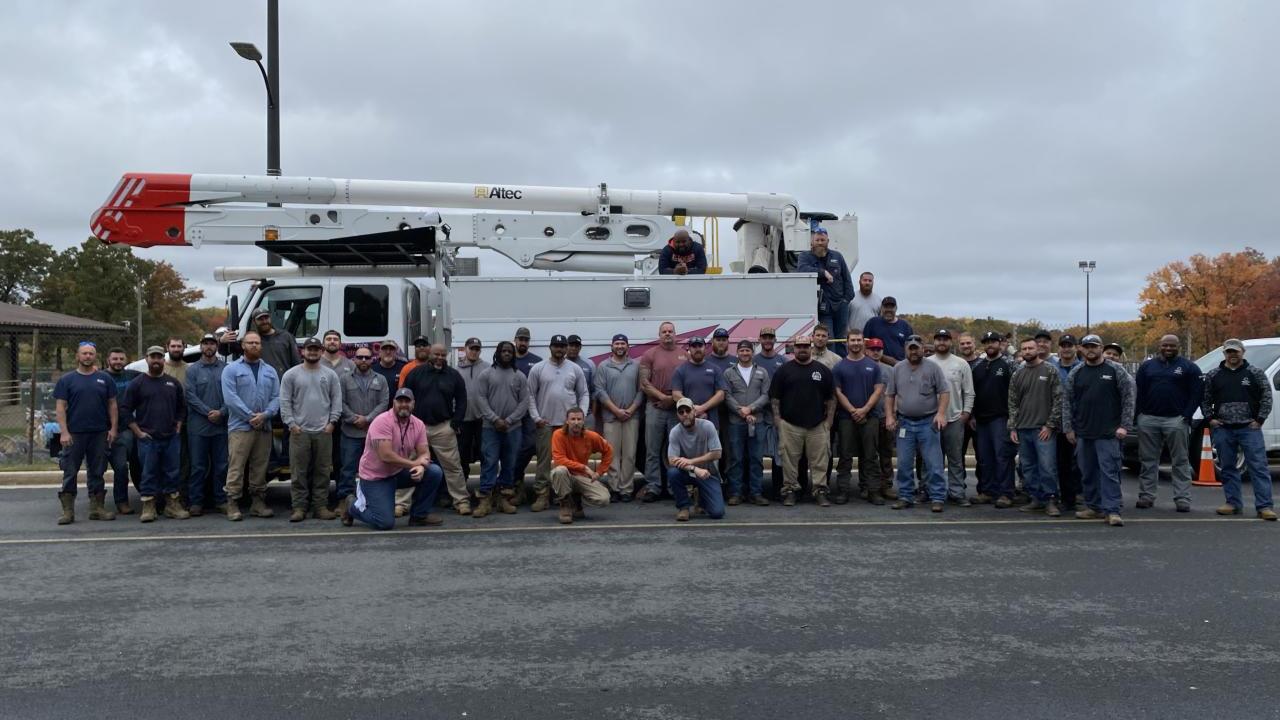 A large group of people posed outside in a parking lot in front of a truck with large extension arm and bucket.