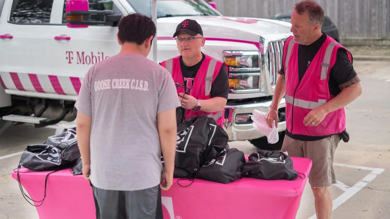 Two people in pink high-vis vests talk to another, a table full of black bags between them. 