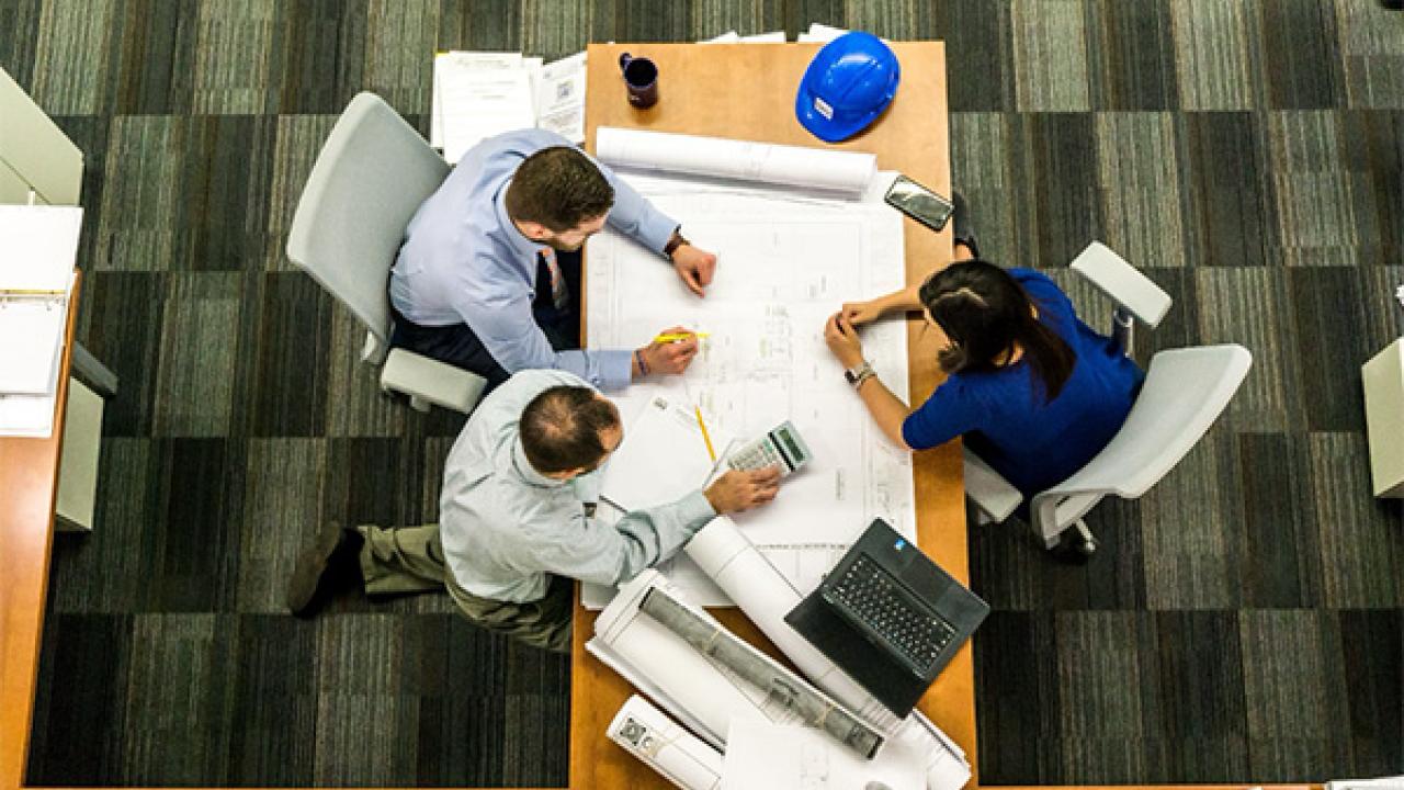 Overhead view of three people looking at blueprints on a shared desk.