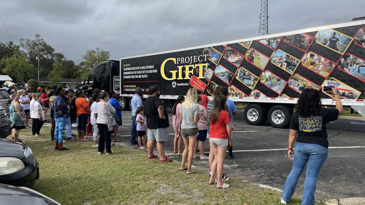 People waiting on a grassy area, looking at a semitruck with "Project Gift" on the side.
