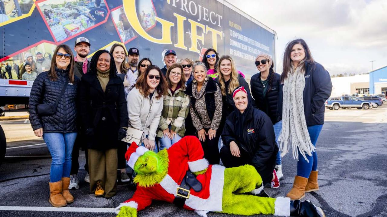 A group posed in front of a semi truck. A person dressed as the Grinch laid out in front of them.