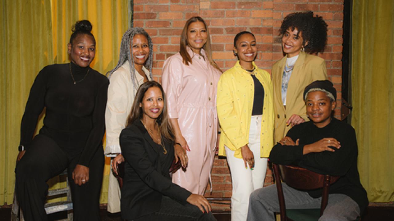 Queen Latifah and a group of others pose in front of a brick wall and yellow curtains