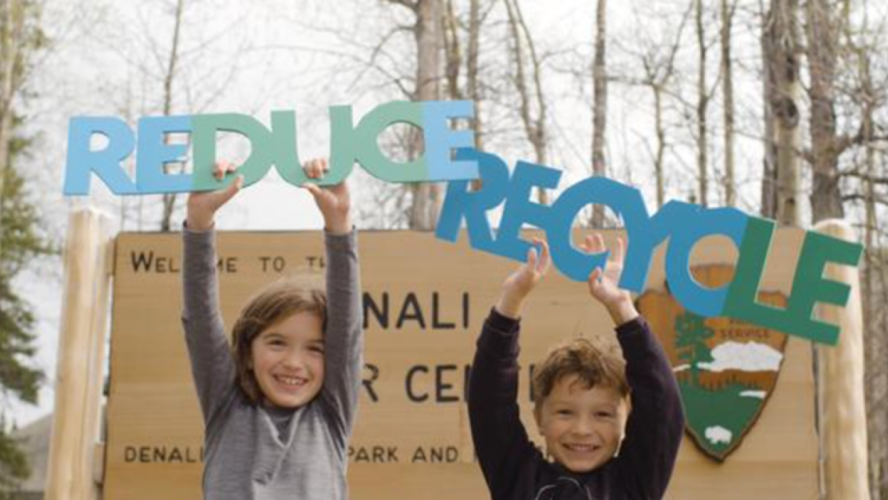 Two children stand in front of a Denali park sign holding up words "Reduce" and "Recycle"