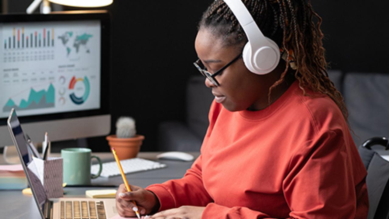 A person with headphones working at a desk by a window with a pencil in hand and an open laptop and separate screen.