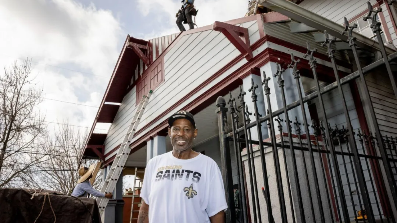 A person in a New Orleans Saints t-shirt standing in front of a house with people doing work on it