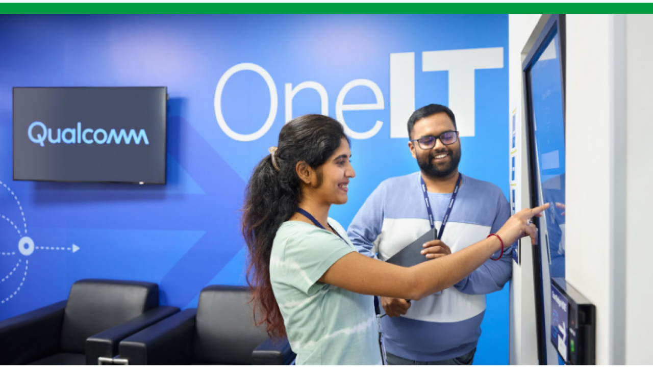 Two smiling people looking at a large touch-screen display. "OneIT" on the wall behind them.