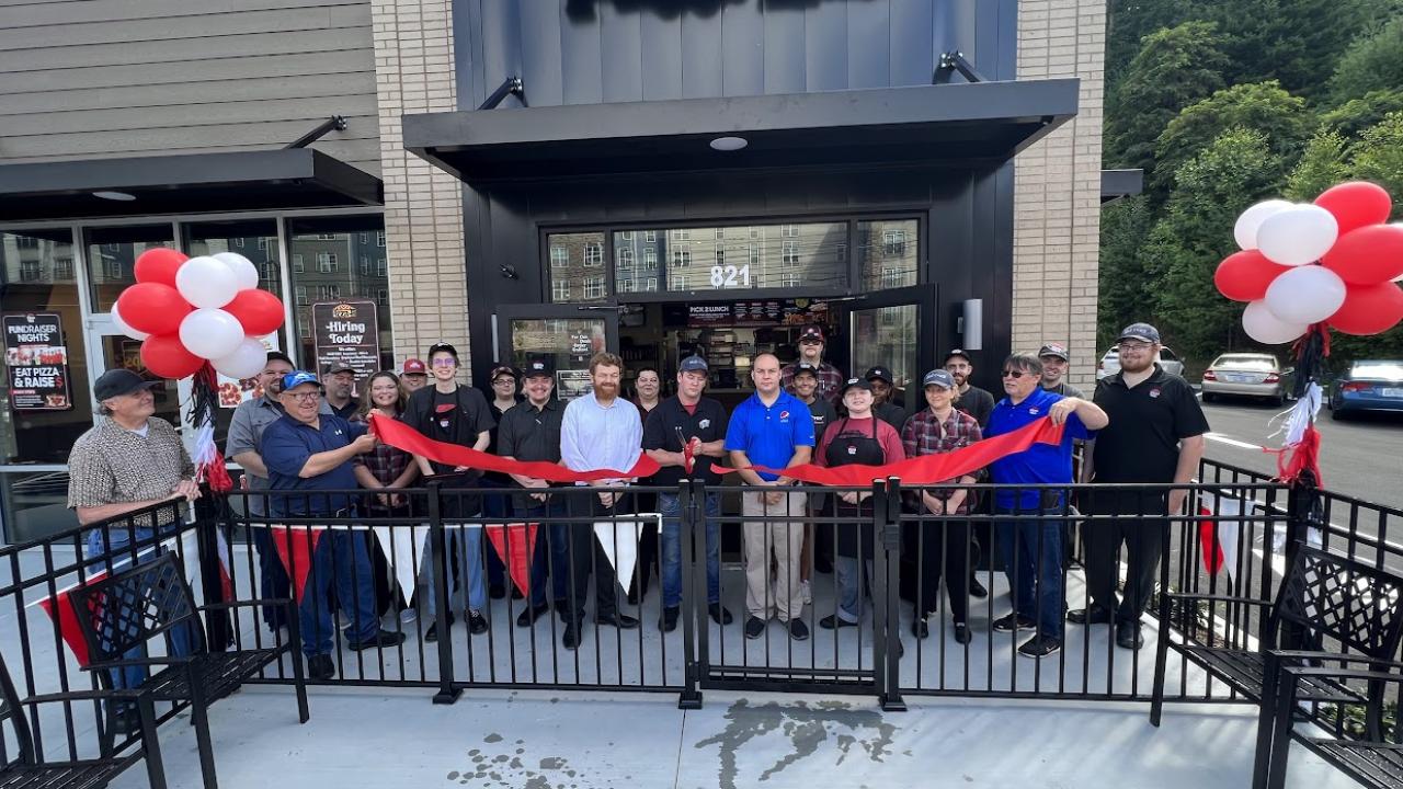 Exterior of pizza hut store, a long ribbon and red and whit balloons. A group of people cutting the ribbon.