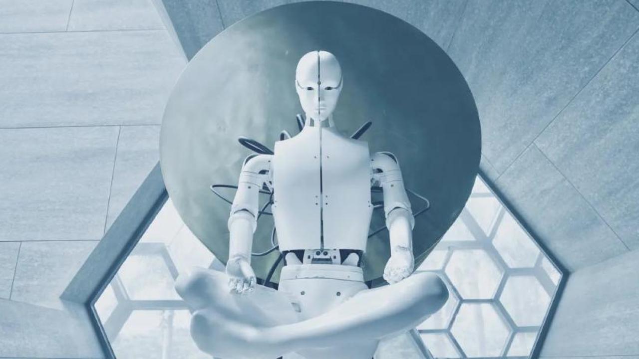 A white human-like robot sitting cross-legged in a stark room of geometric shaped windows and walls.