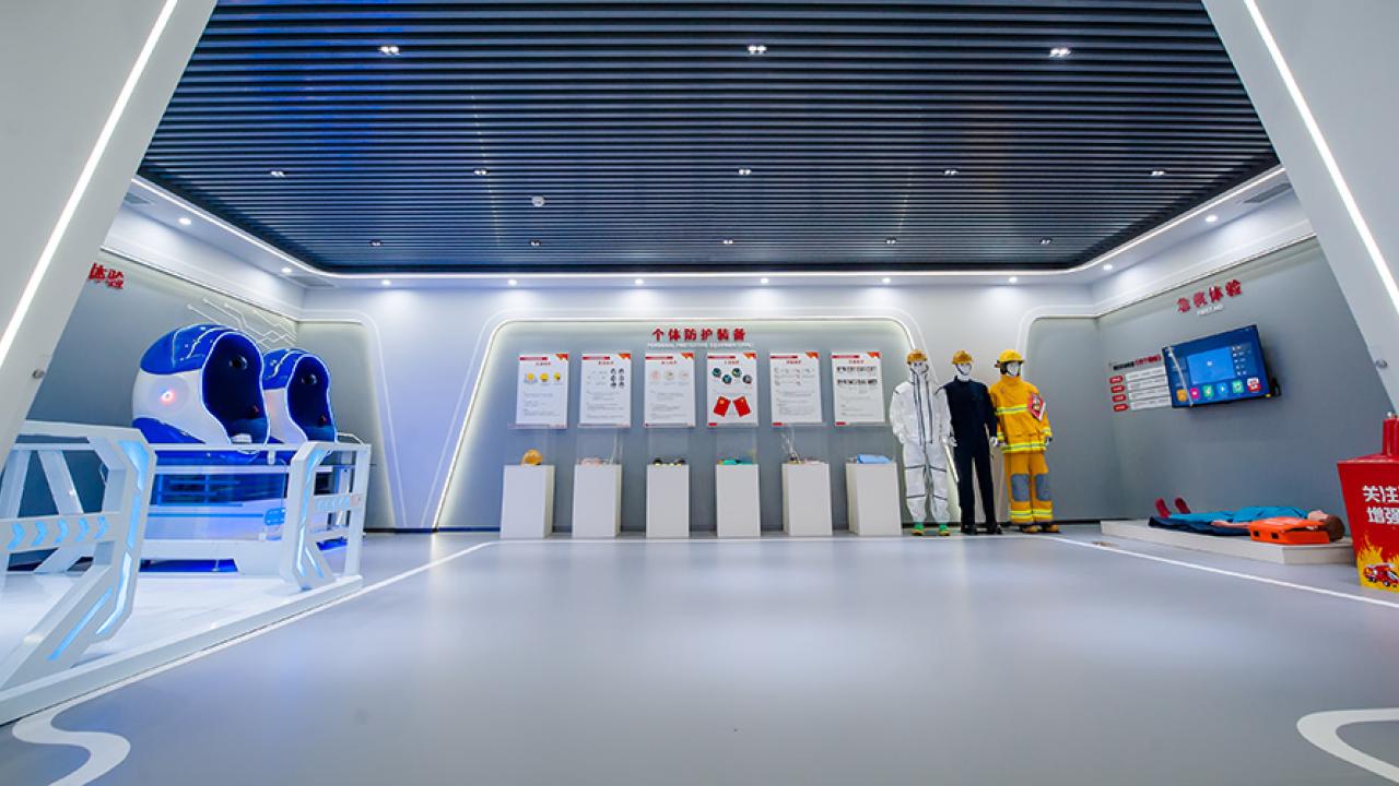 A futuristic looking room with dummies in safety garments, posters, and another dummy on a mat on the floor against the right wall next to a box with flames on it. A monitor above the dummy on the wall. Two pods on the left wall. 