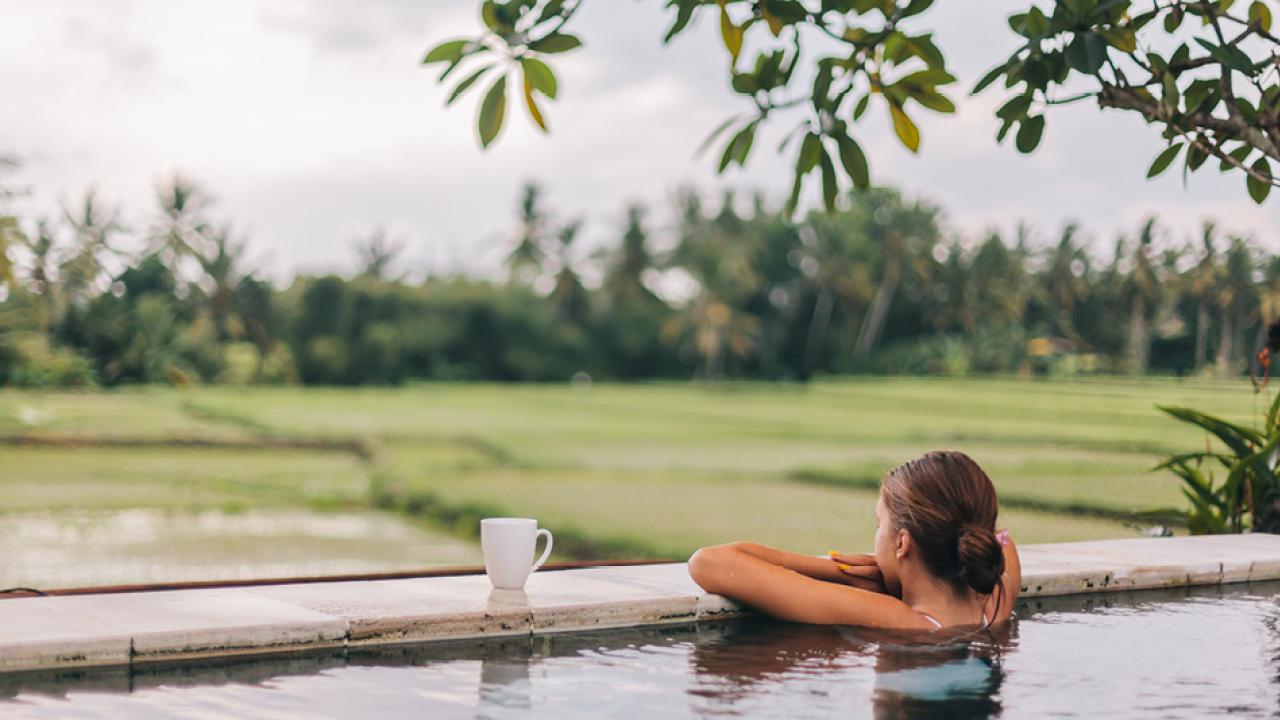 A person in an outdoor pool, arms crossed over the ledge overlooking a field and trees.