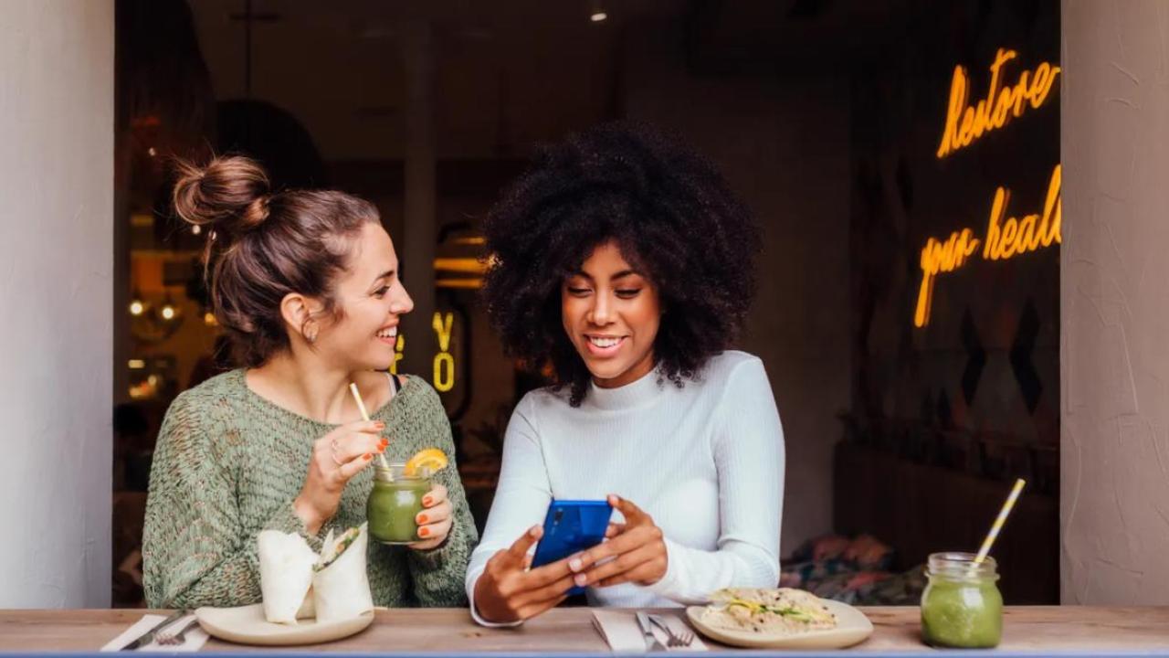 Two smiling people seated with food and drinks. One showing the other their phone.
