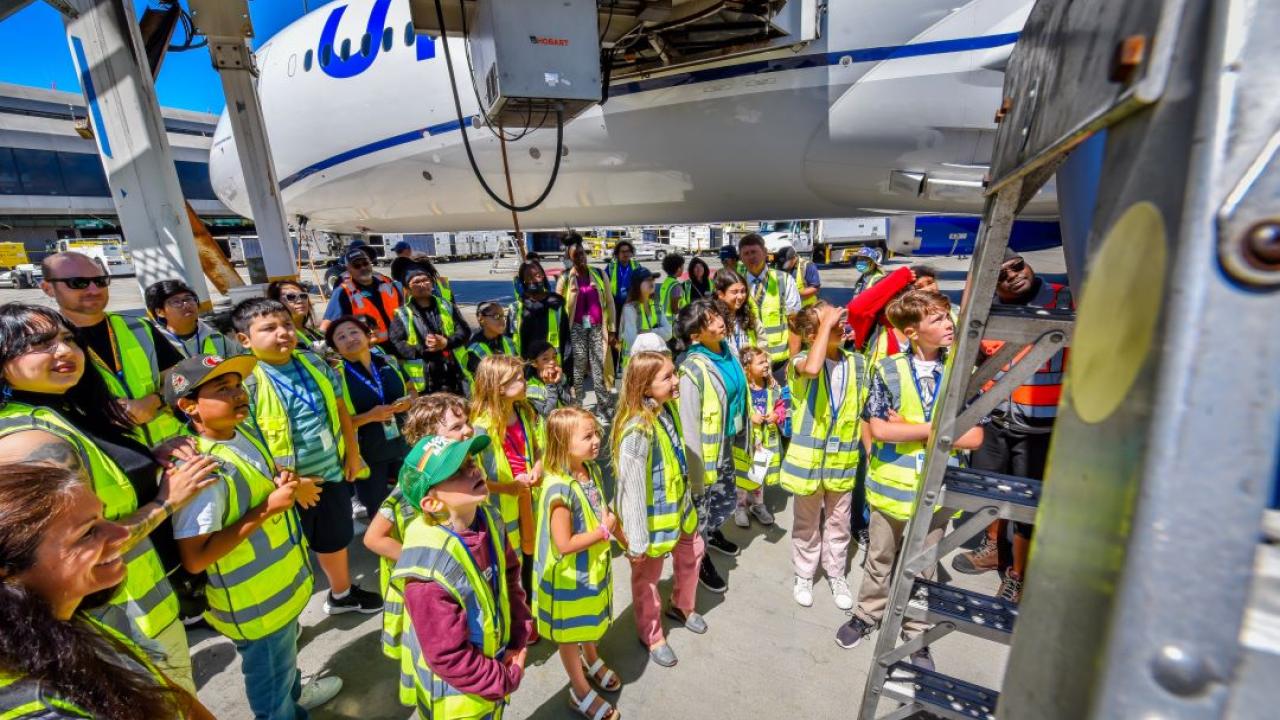 A group of kids and adults in high-vis vests looking up close at an airplane.