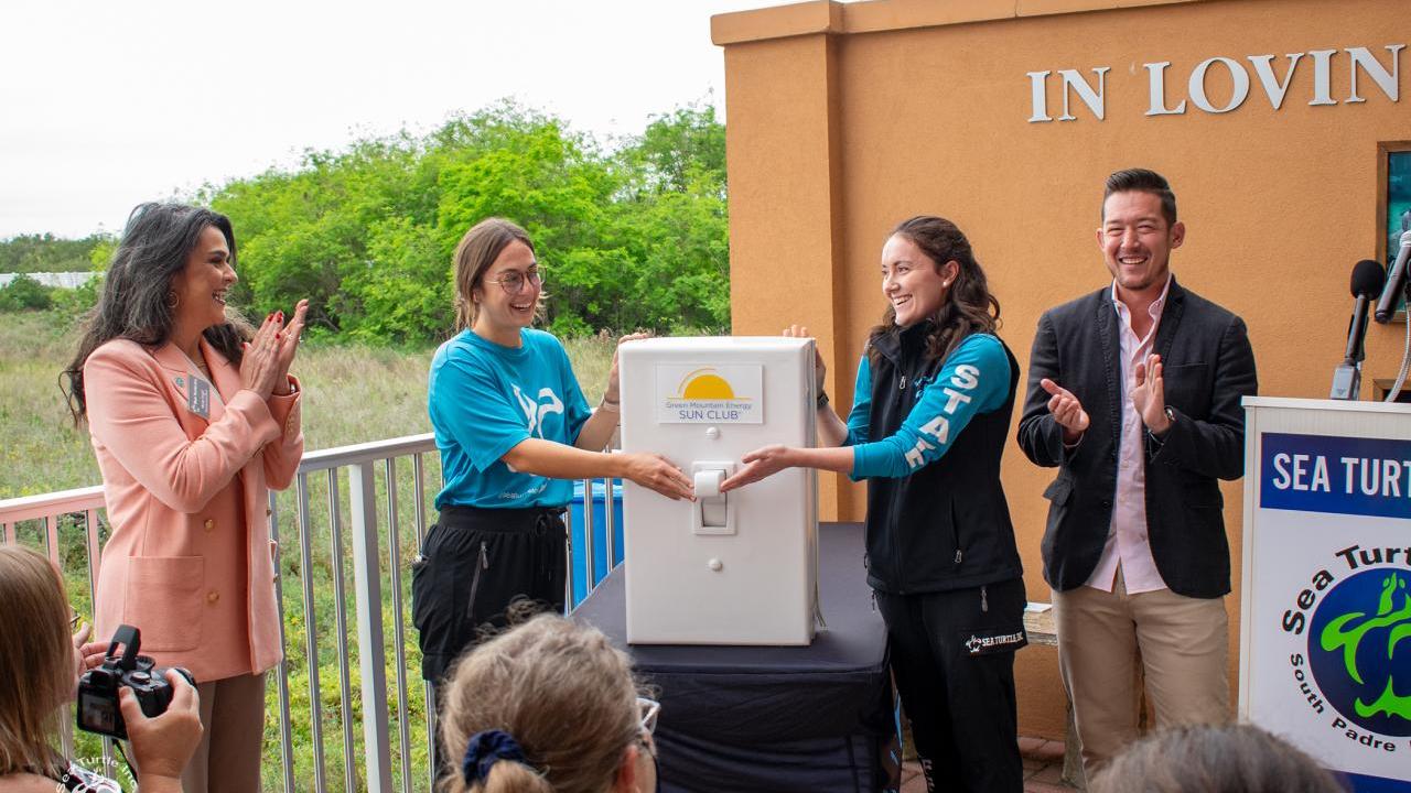 Two women in blue shirts flip an oversized prop light switch as onlookers smile and clap