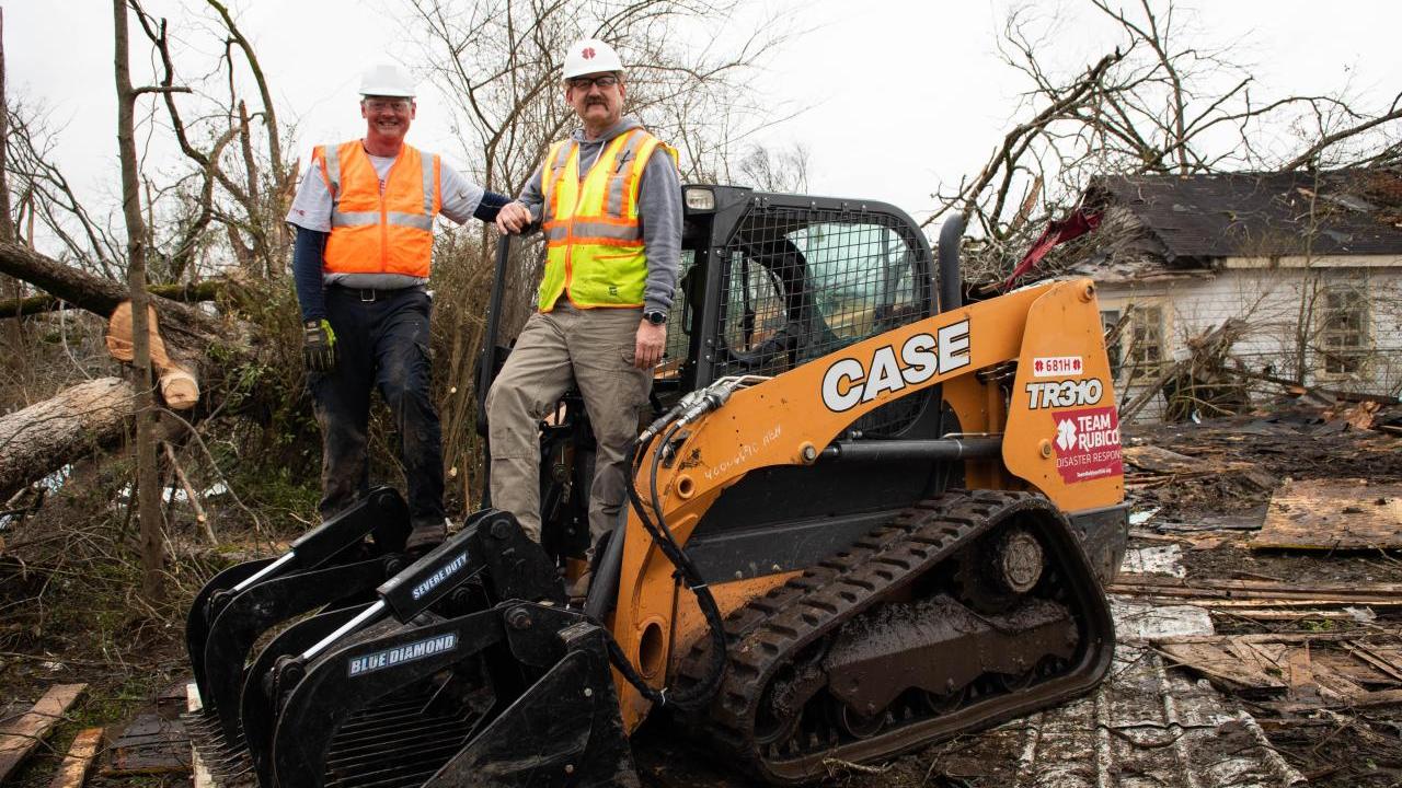 Two people in safety vests and hats standing on a CASE front-loader. Debris all around them.