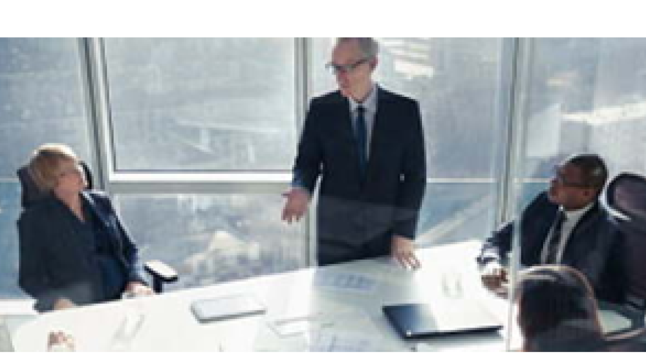 One person standing in a conference room in a tall building, others seated at the table.