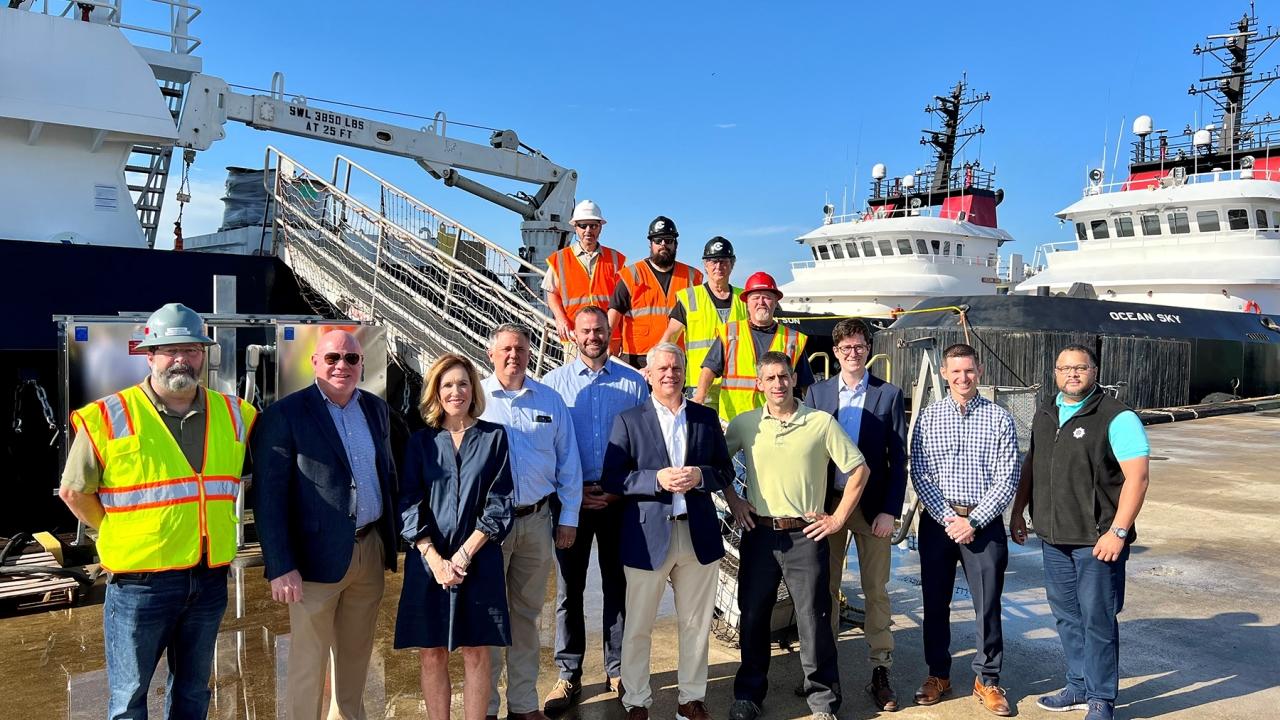 A large group of people standing on a pier in front of a large ship. Some in business attire and some in high-vis and protective wear