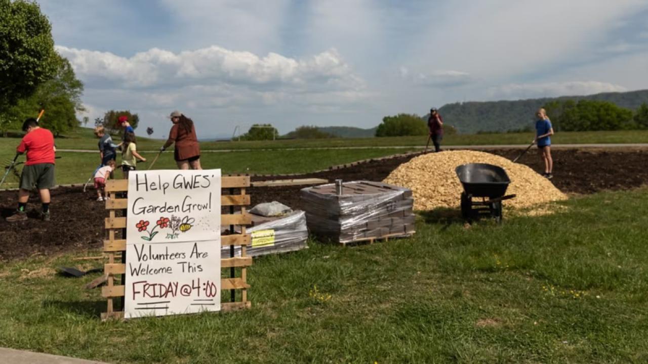 Volunteers working outside with a sign on wooden pallets that reads "Help GWES' Garden Grow Volunteers Are Welcome This FRIDAY at 4:00"
