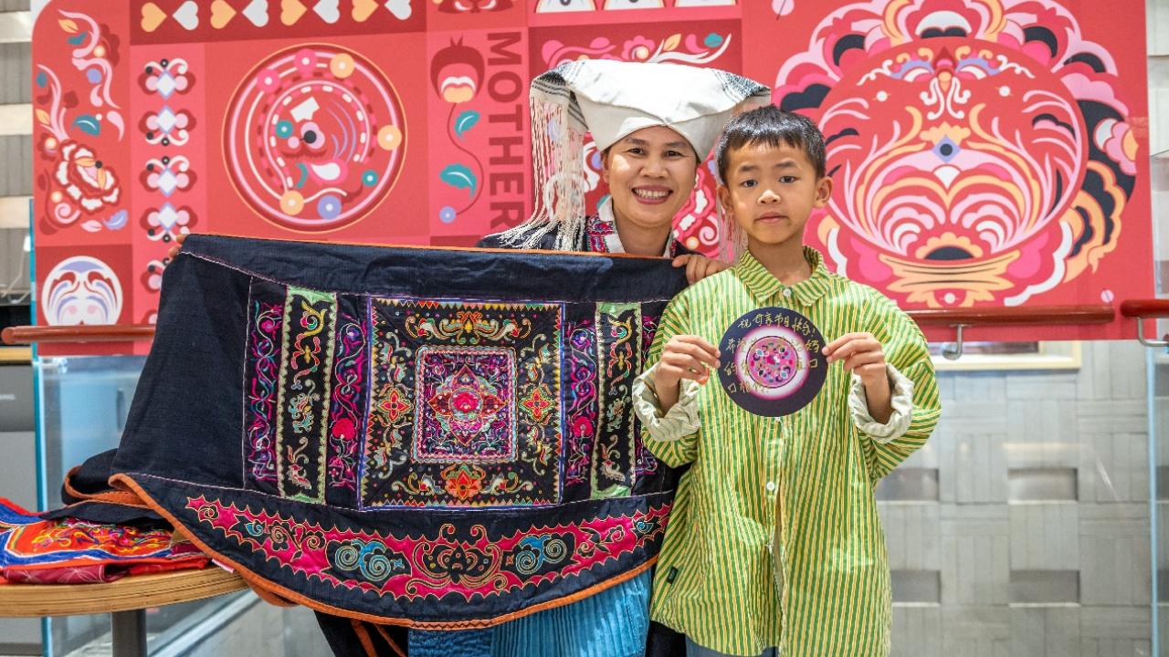 a child stands beside Mrs. Huang who is holding an embroidered sling. Cultural art behind them
