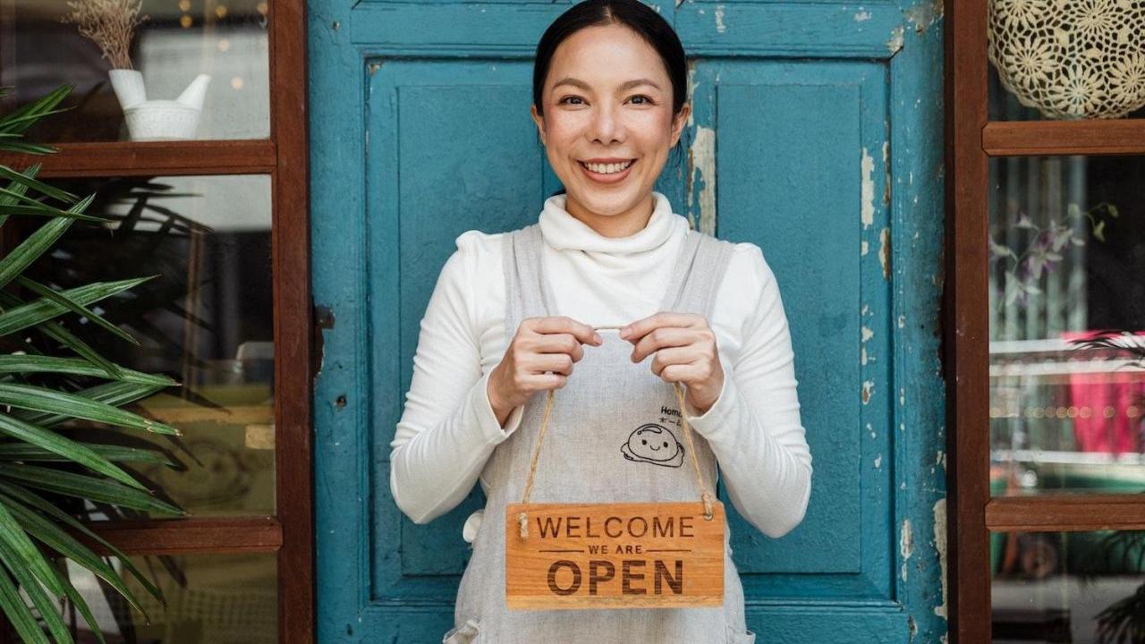 A smiling person holding a wooden "Welcome we are Open" sign in front of a blue door.