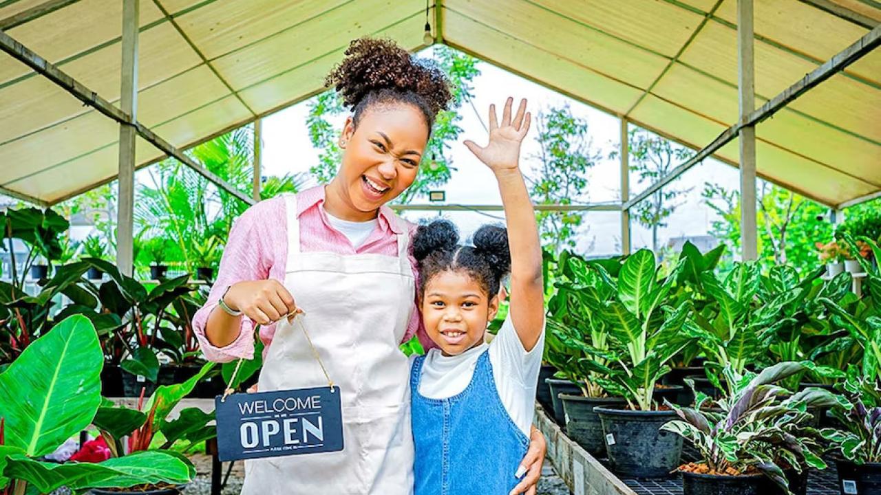 An adult and child standing in a green house, one holds an Open sign.