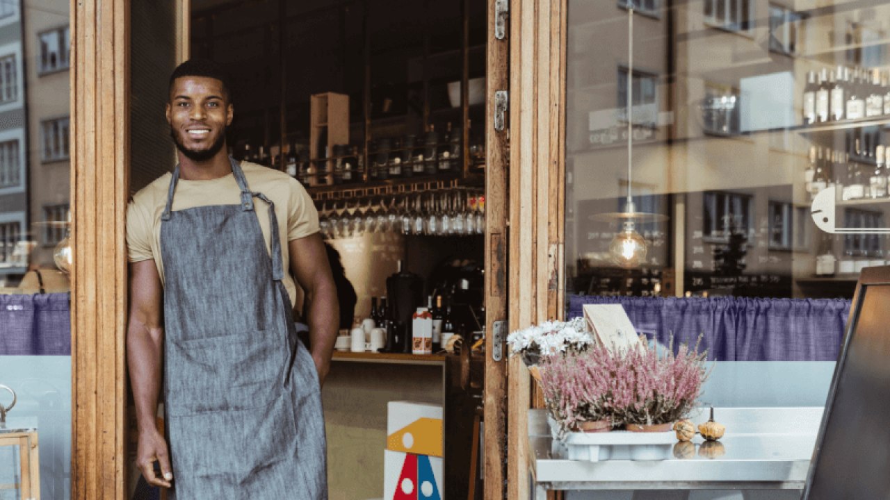 A smiling person wearing an apron, leaning against the door frame of a business.