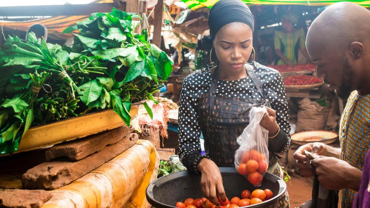 A person bagging produce for another watching on.