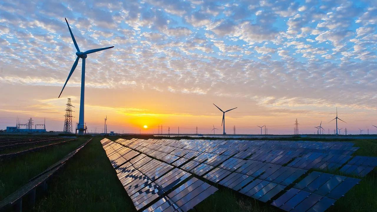 Rows of solar panels with wind turbines and a setting sun in the distance. 