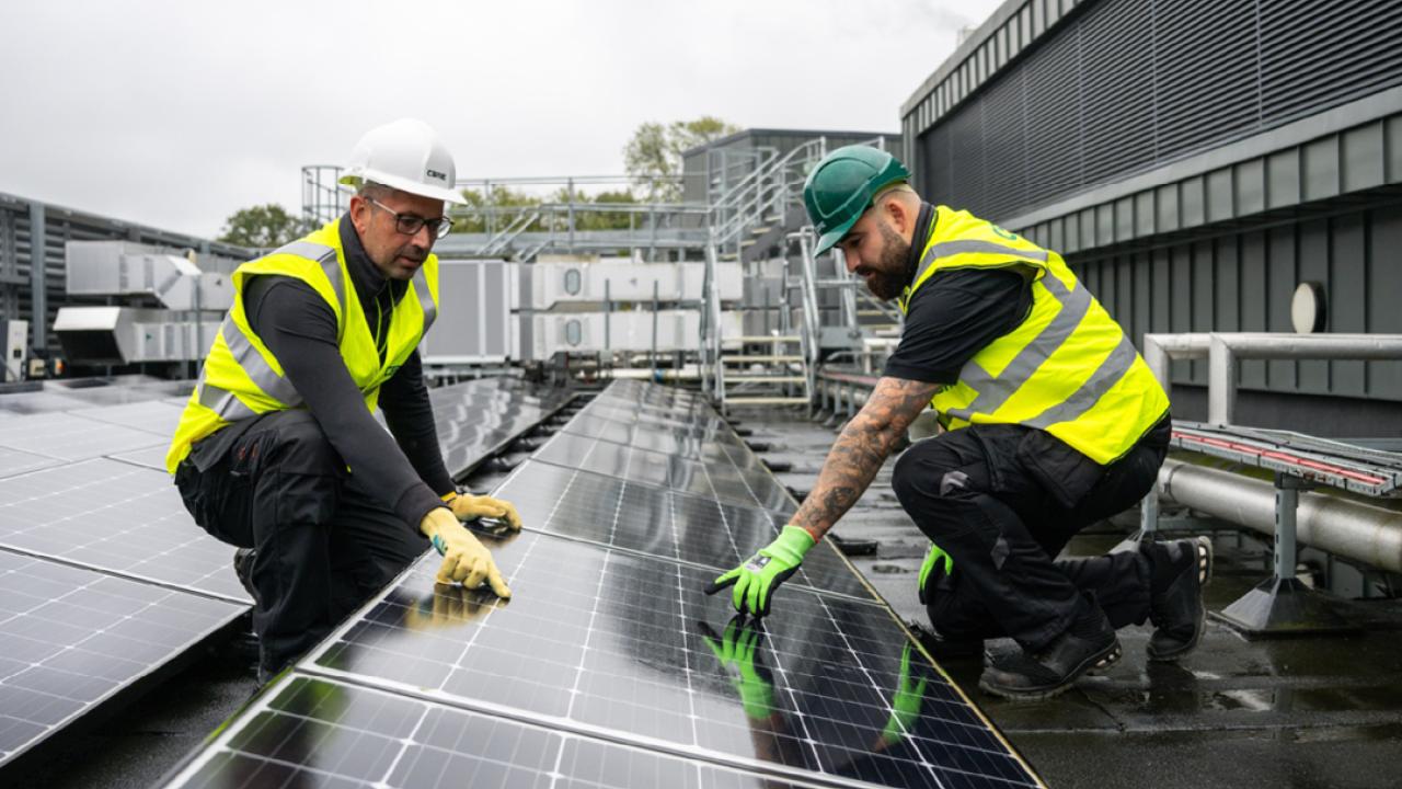 Two people in high-vis vests and hard hats pointing to a solar panel in a row of others outside.