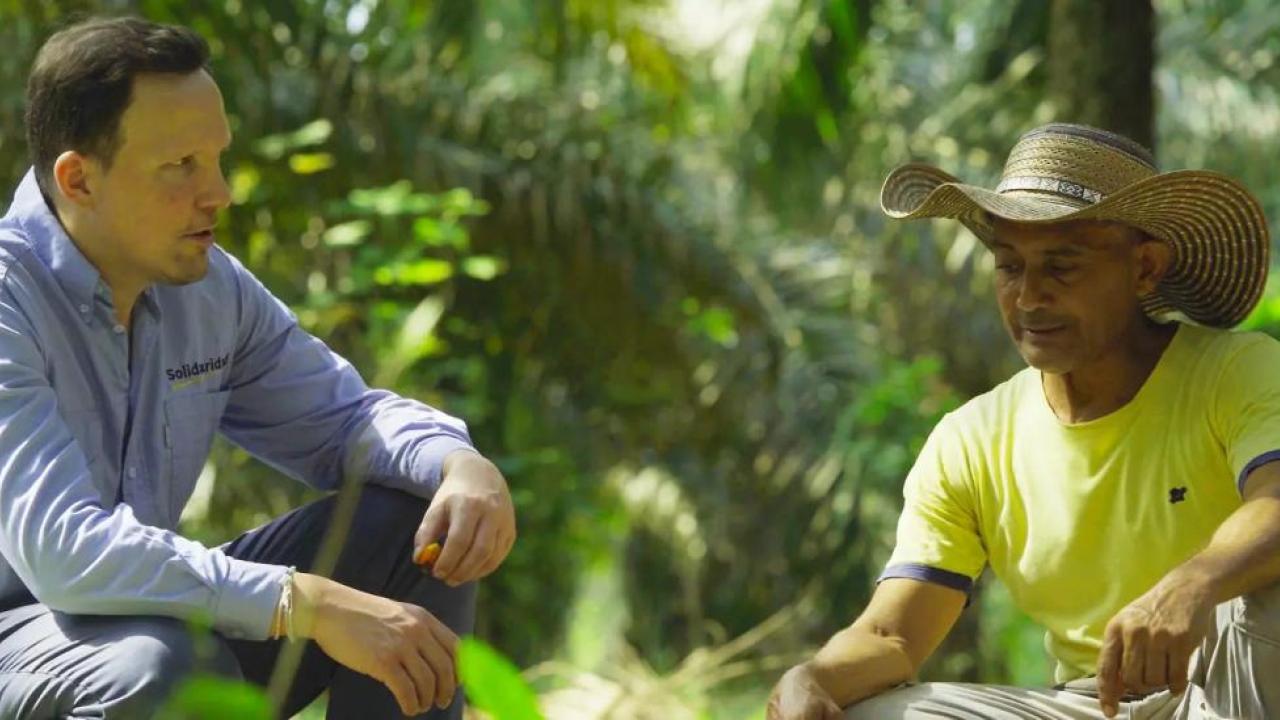 Two people crouched, talking together, in a jungle setting.