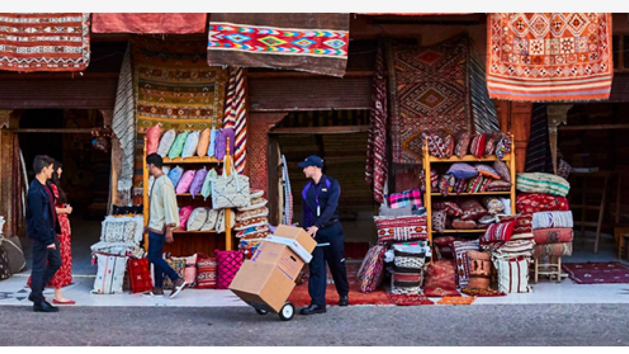 A person delivering boxes at a shop with colorful textiles hanging.