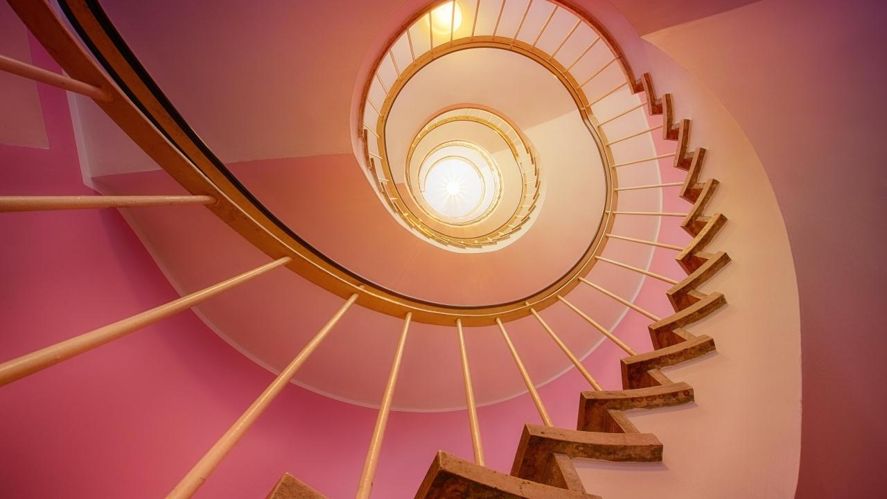 Looking up a long spiral staircase, pink hues on the wall.