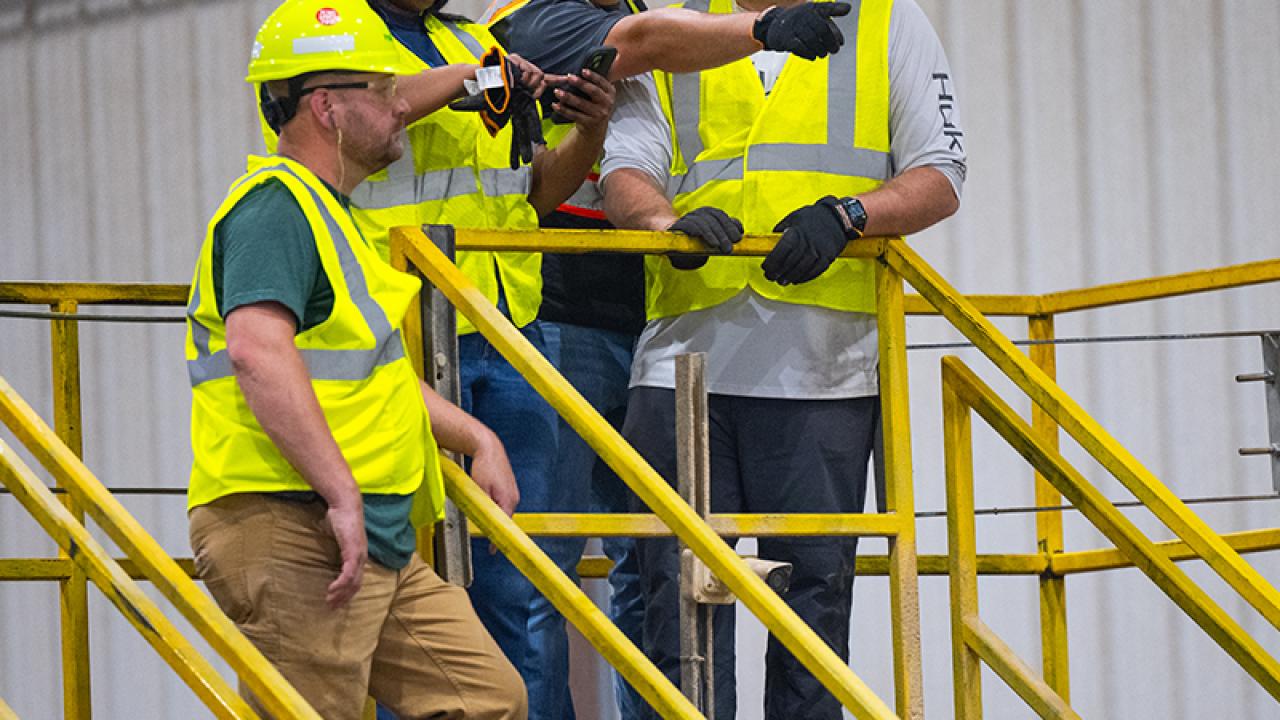 Four employees in hard hats and high-vis vests standing on a platform. One pointing to the distance.