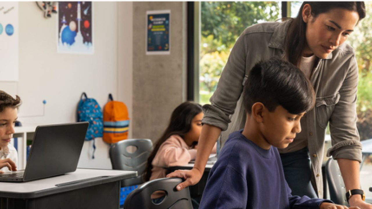 a teacher assists a student on a laptop, two other students at desks behind them