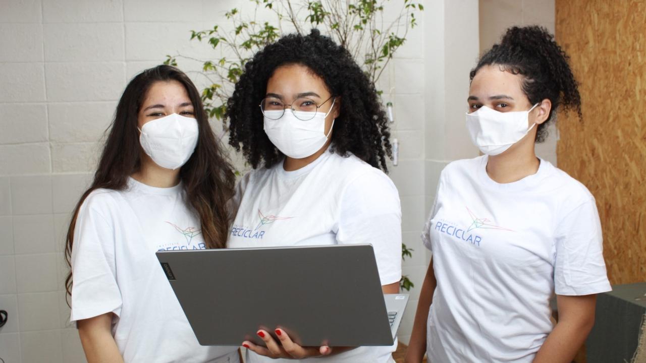 Three students wearing the same "Reciclar" t-shirts and protective masks. The middle student holding a Lenovo laptop.