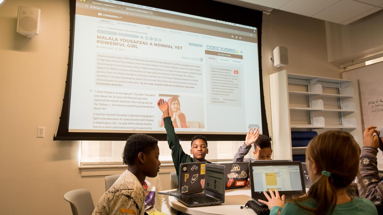 students at a round table, each on a laptop. A large pull-down projection screen behind them with a presentation on it.