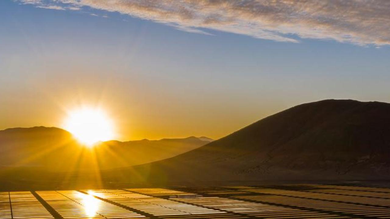 Panoramic view of hilly landscape, a setting sun in the distance and a large field of solar panels.