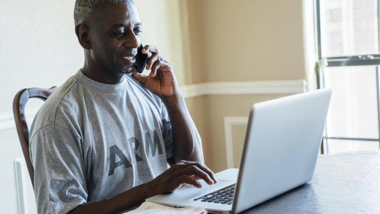 A person in an Army t-shirt on the phone while typing on a laptop