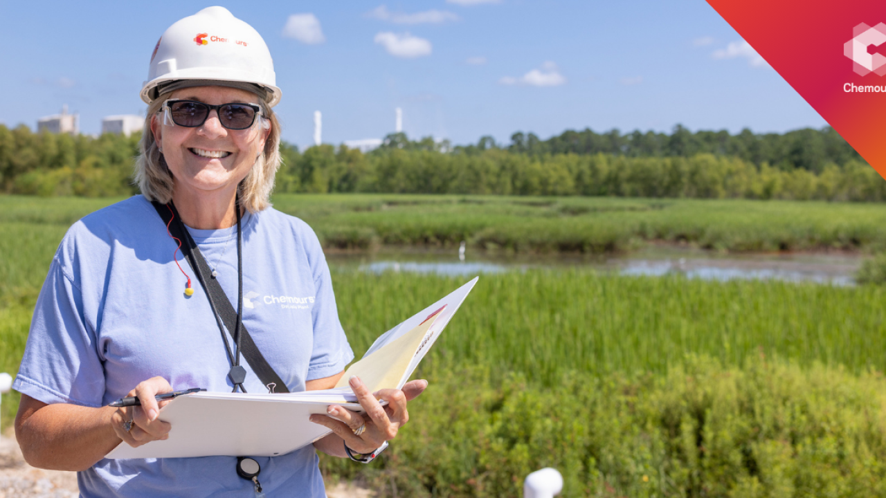 A person smiling outside, holding a notebook and wearing a hard hat