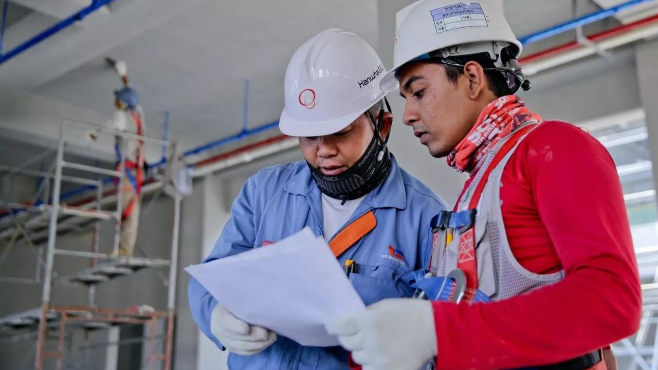 Two people in hard hats, gloves and protective masks looking at a piece of paper. A person standing on scaffolding painting the ceiling behind them.