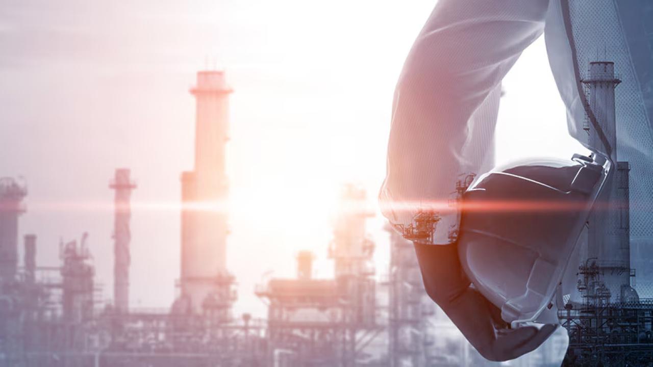 A person holding a hard hat, smoke stacks in the distance.