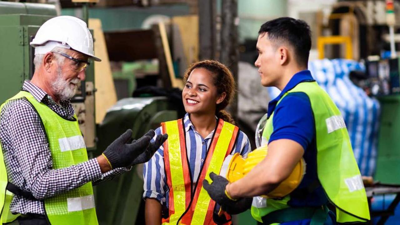 Two workers watch as a third talks with them. In an industrial setting, all are wearing high-vis vests.