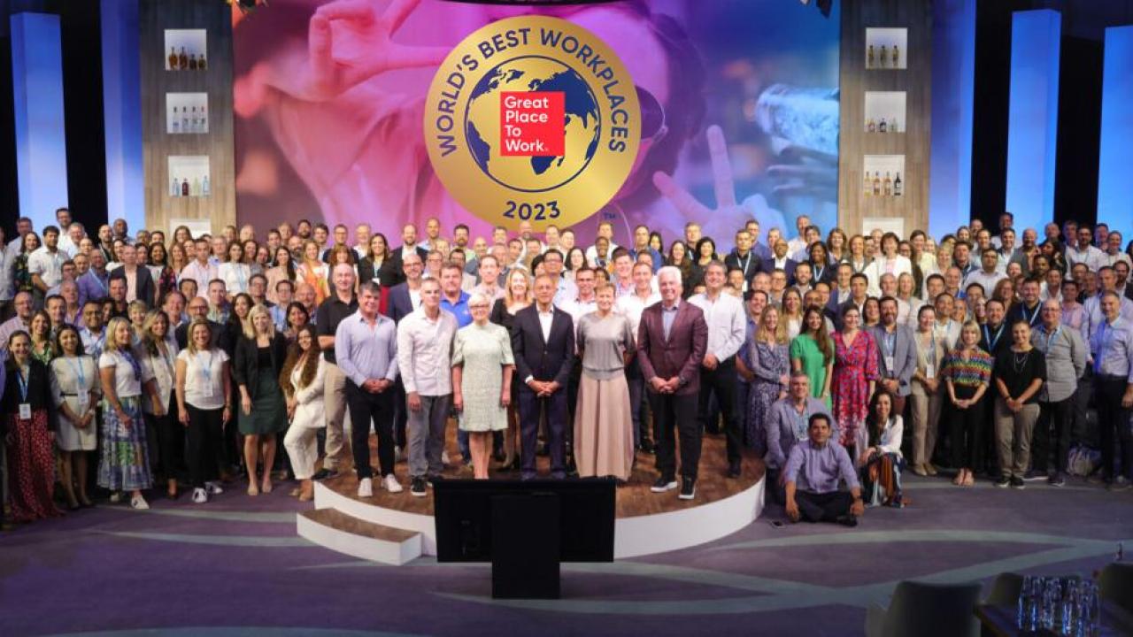 A large group posed on a stage. "World's Best Workplaces" banner above them.