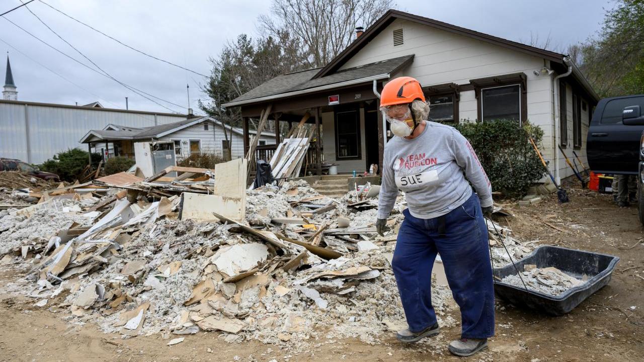A volunteer in hard hat and respirator pulling a black sled full of rubble. A pile of rubble in front of the house behind them.