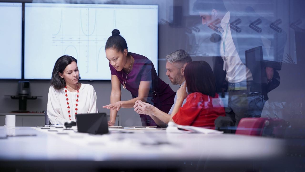 five people in a conference room, two standing and all looking at the same screen.