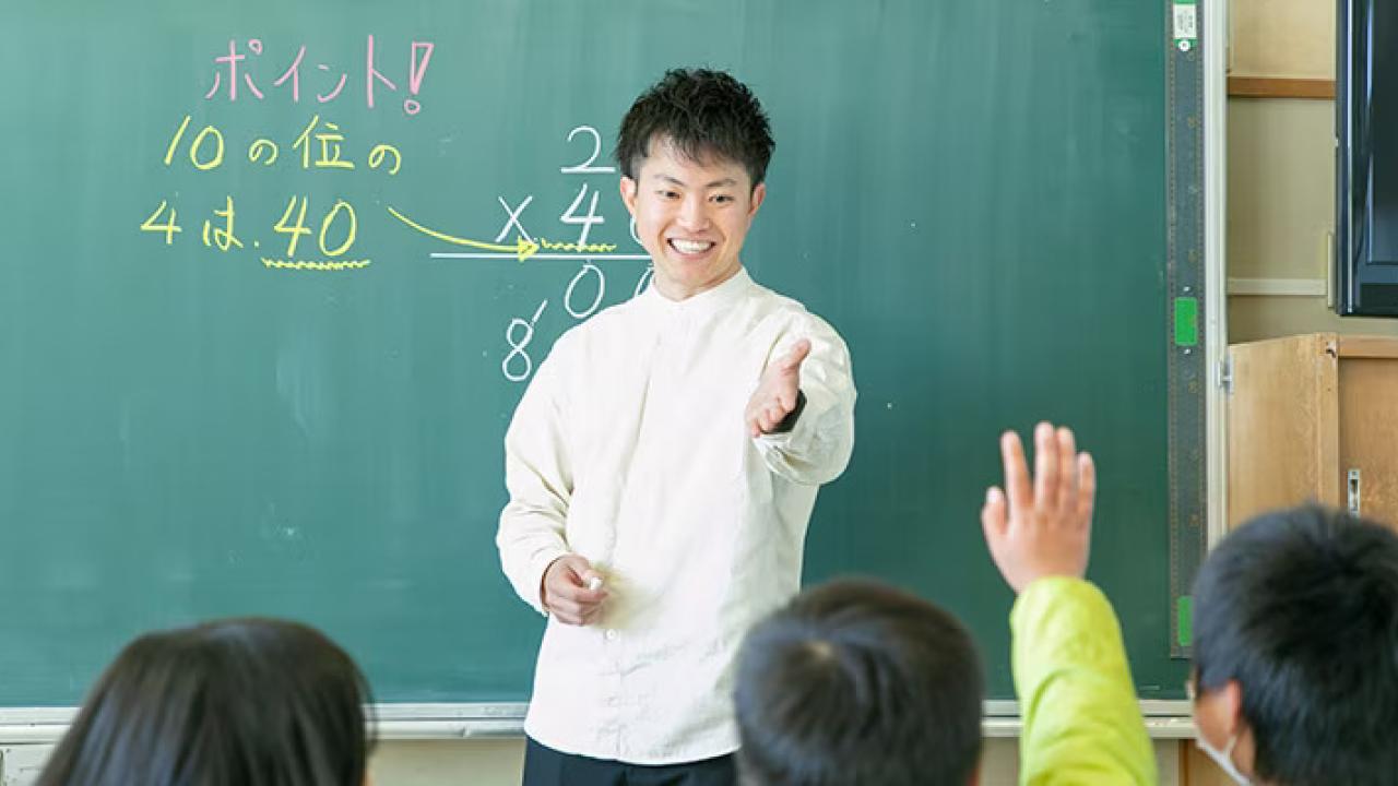 A teacher in front of a chalkboard, pointing to a student.
