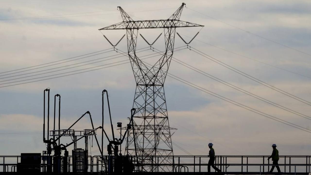 silhouettes of two people on a walkway leading to industrial electricity equipment. A tall power station and tower in the background.