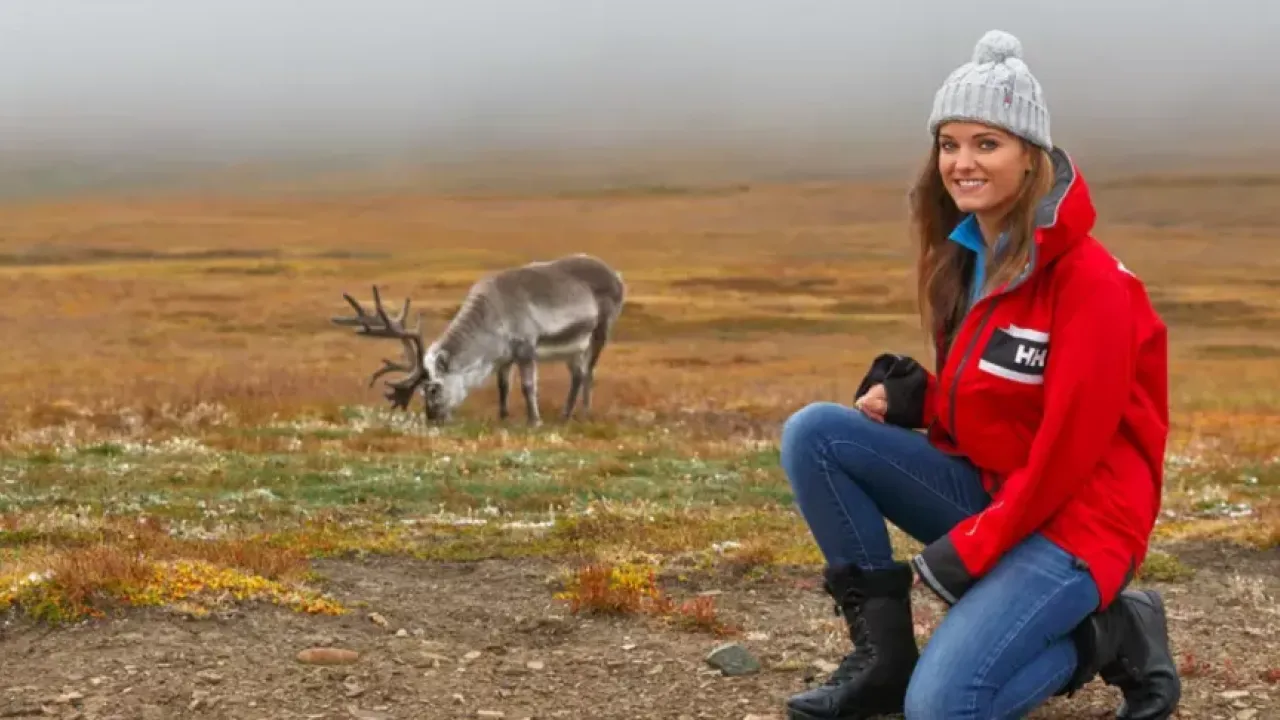 A person crouched on an open plain, a caribou in the distance
